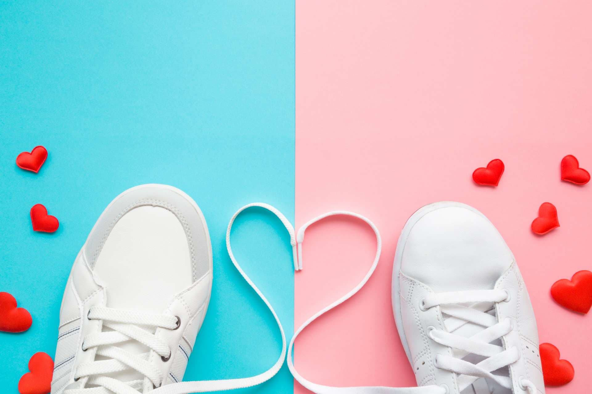 Herz aus weißen Schnürsenkeln zwischen männlichen und weiblichen Sportschuhen. Helle, rote Herzen. Liebeskonzept. Leerer Platz für schönen, niedlichen Text, Zitat oder Sprüche auf pastellblauem und rosa Papier. Nahansicht.