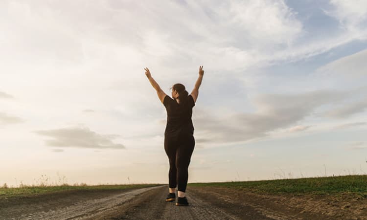 Übergewichtige Frau auf Landweg bei Sonnenaufgang, die aufsteigende Hände zum Himmel feiert