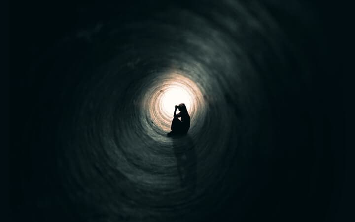 The silhouette of a praying woman sitting in a dark tunnel with a light at the end.