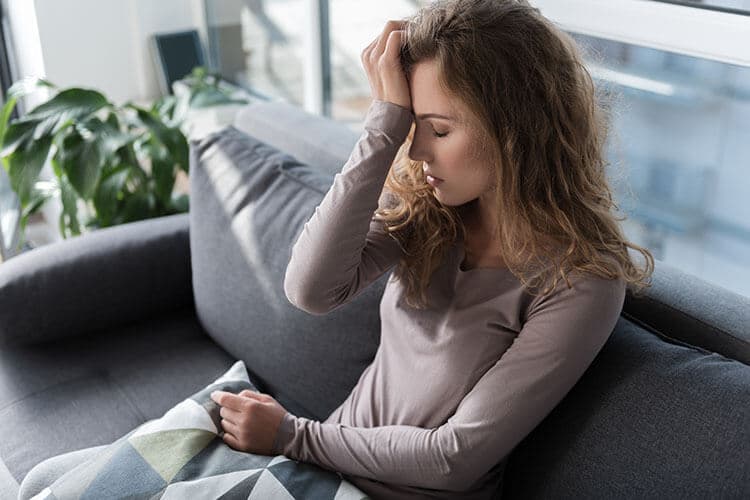 Junge Frau mit Migräne sitz zuhause auf dem Sofa und hält sich den Kopf.