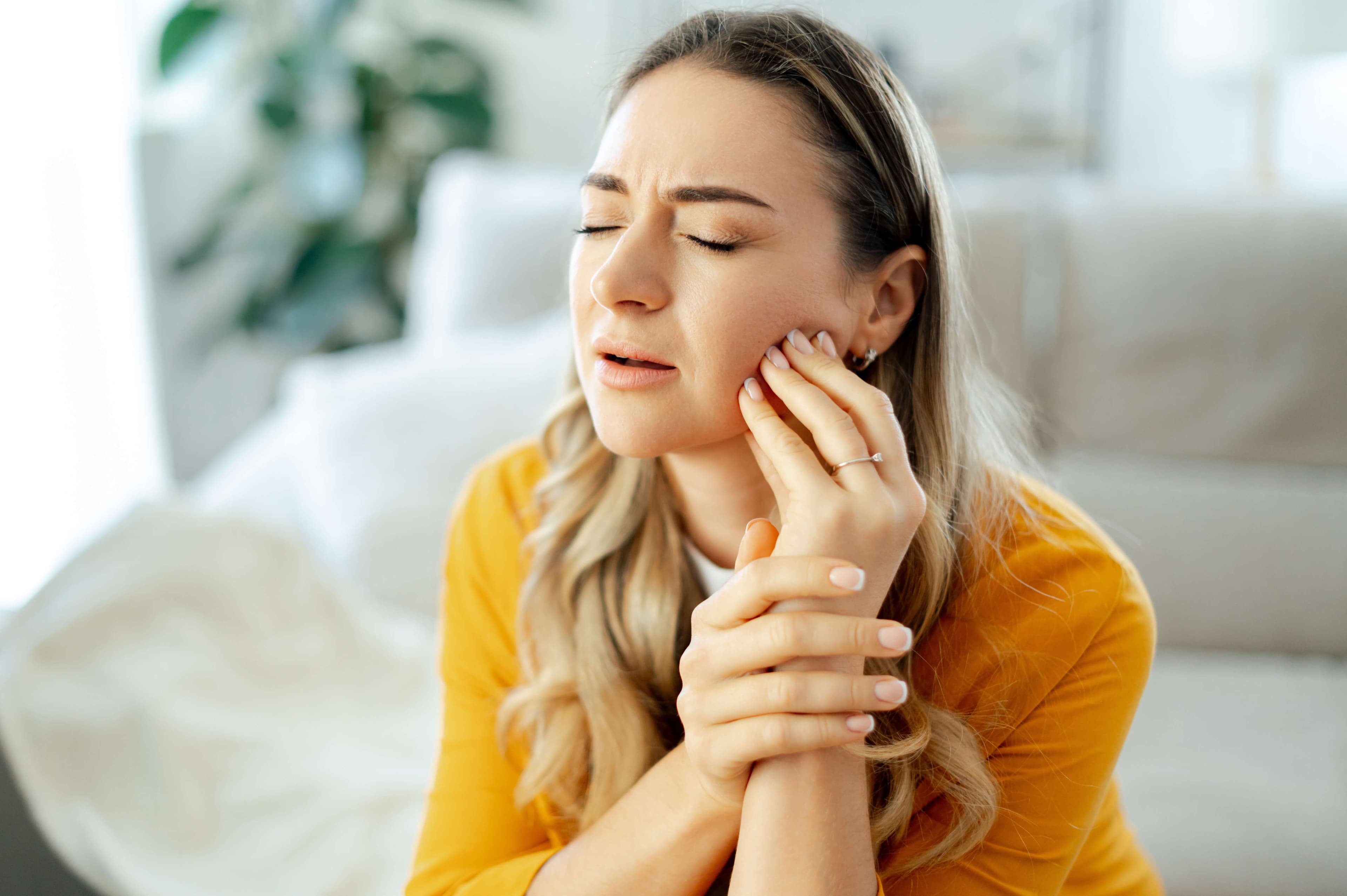Upset caucasian woman in a yellow shirt sitting on a couch at home with eyes closed, holding her cheek in pain with a pained