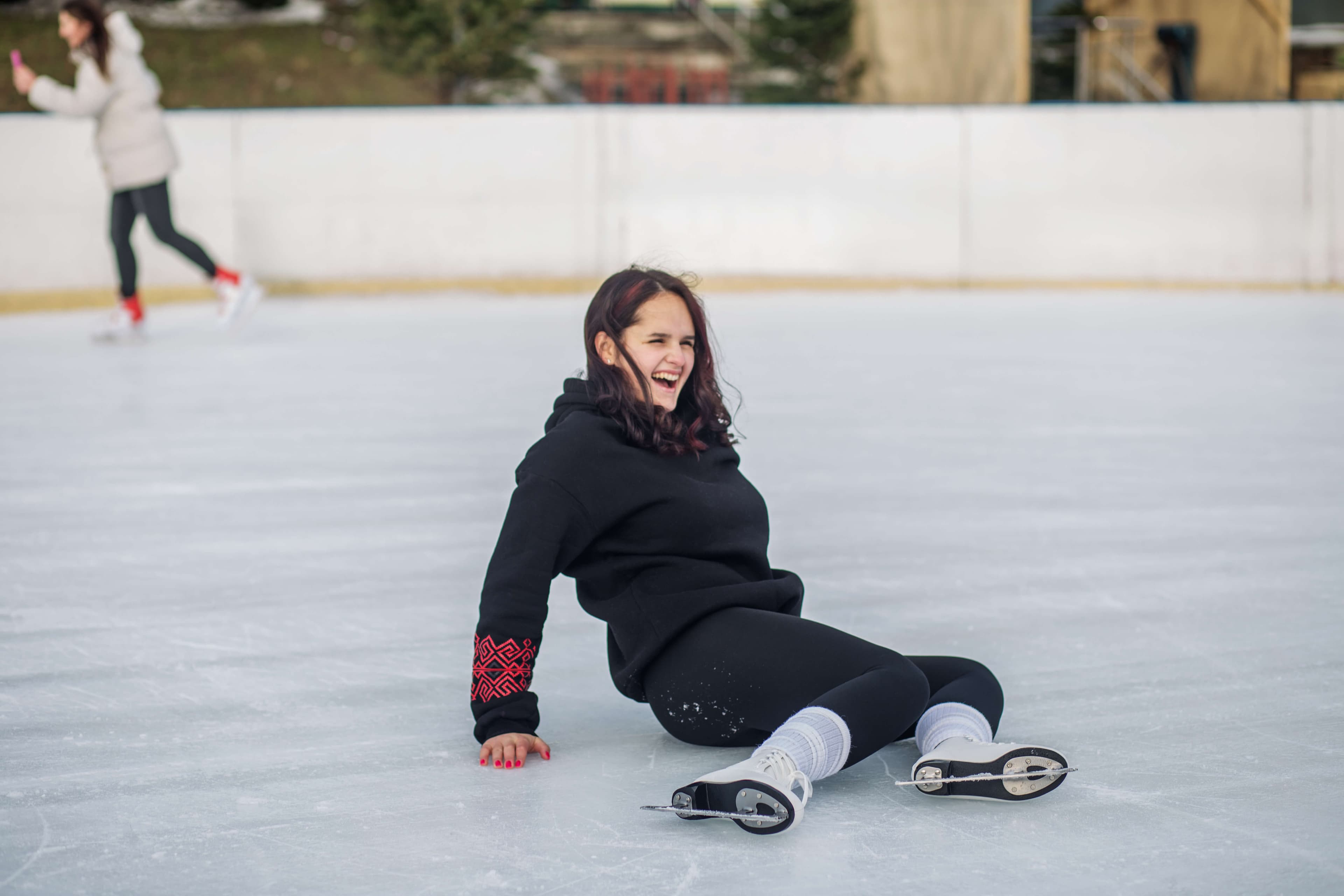Teenage girl sits on an outdoor ice rink, laughing joyfully after a fall, enjoying a fun winter skating experience.