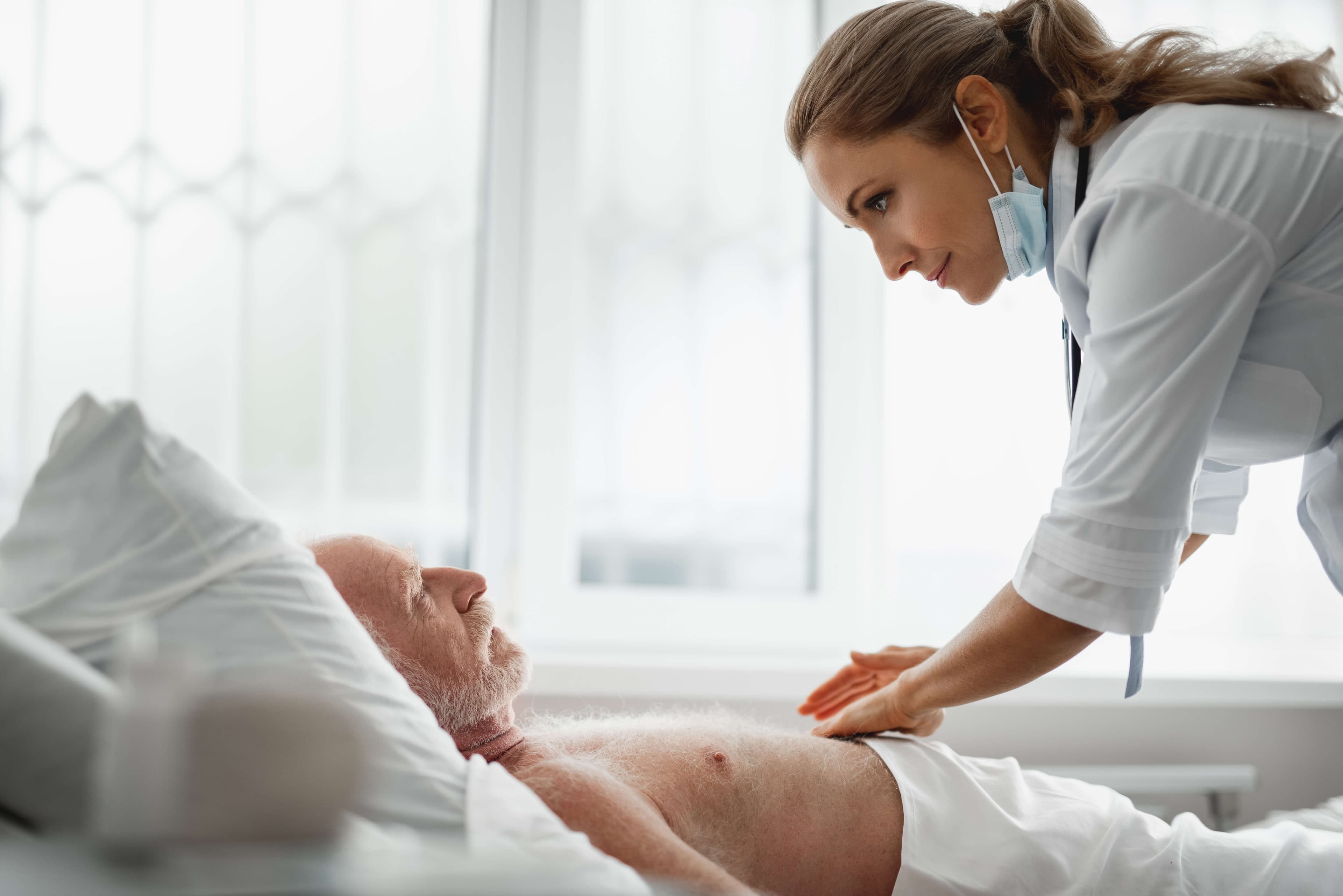 Side view portrait of shirtless old man lying in bed while physician palpating his abdomen. Large window on background