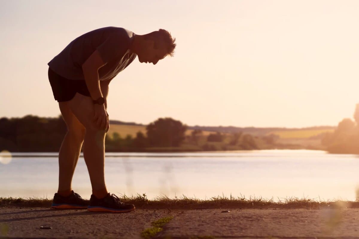 Un jeune homme fait son jogging au coucher du soleil au bord du lac ; l'athlète fatigué se tient le genou douloureux.