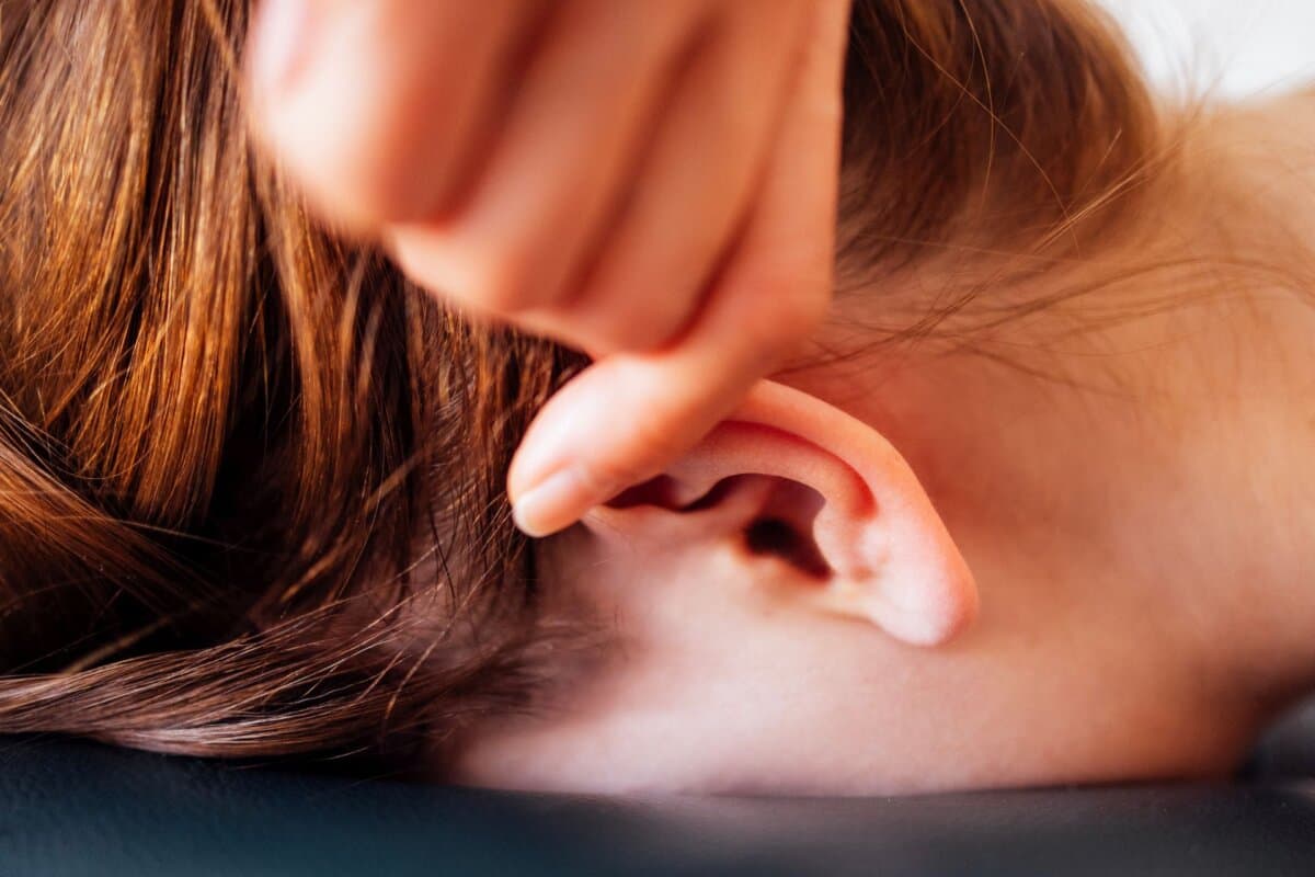 Detailaufnahme einer Ohrakupressur: Die Hand der Therapeutin am Ohr, das Gesicht der Patientin entspannt hinter der