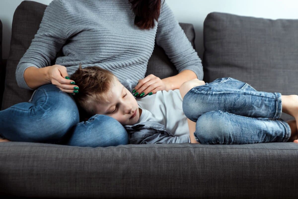 Junge liegt mit Bauchschmerzen auf dem Sofa, seine Mutter ist besorgt an seiner Seite.
