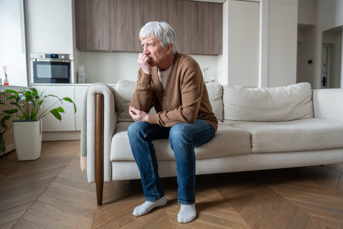 Ein älterer Mann sitzt sichtlich belastet auf einem Sofa.
