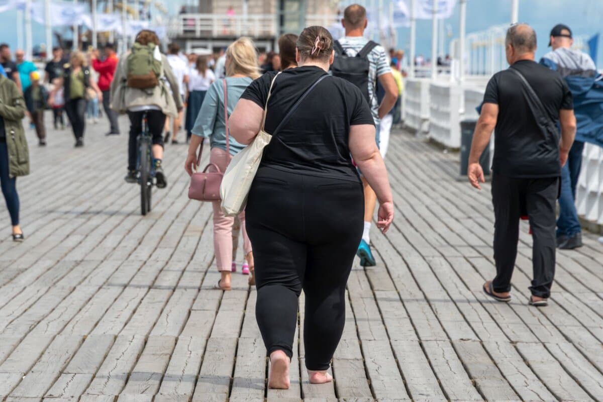 Übergewichtige Frau geht barfuß auf einem Pier zwischen normalgewichtigen Menschen.