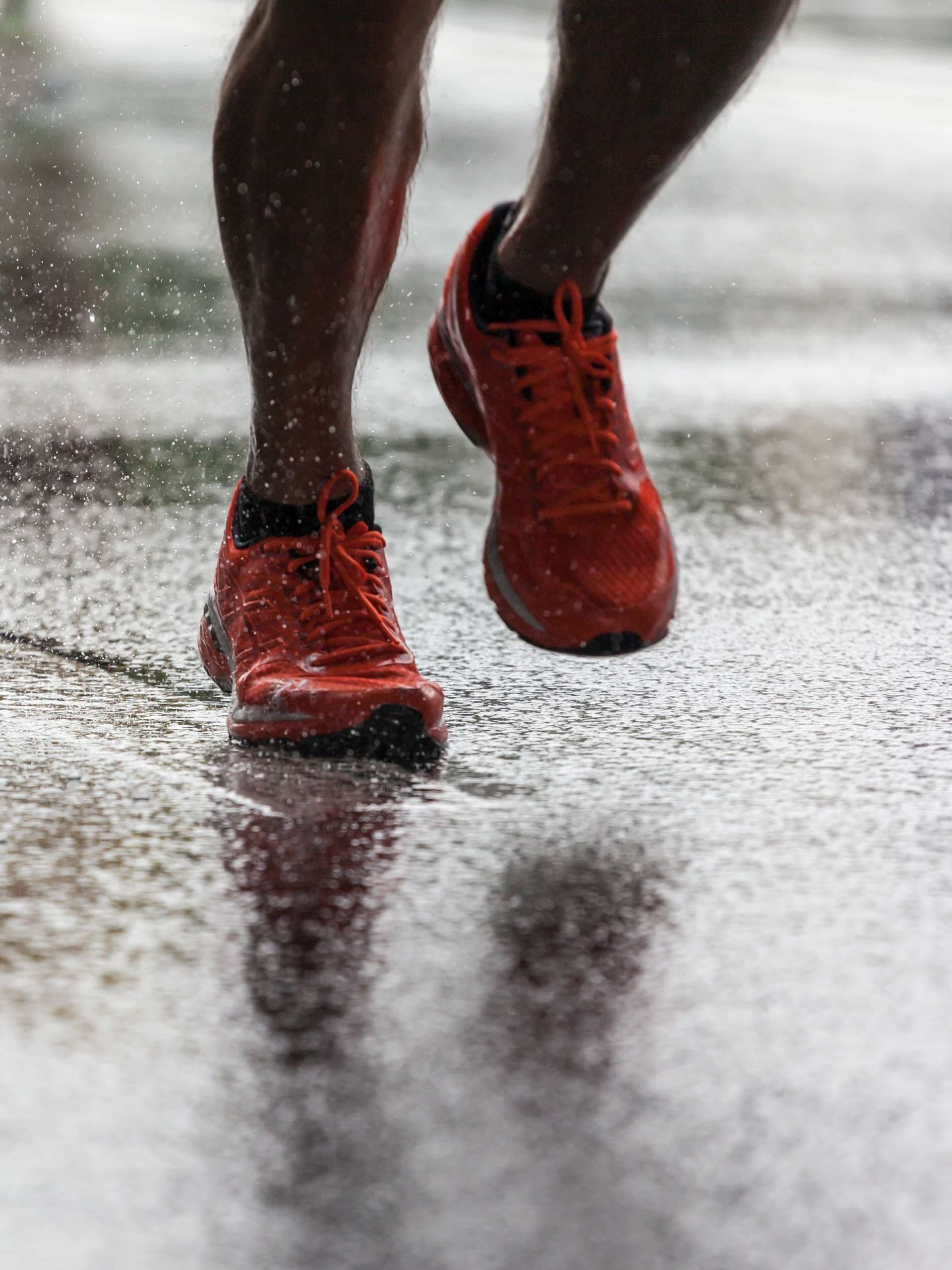 Nahaufnahme von Sportschuhen eines Athleten, der auf einem nassen Asphalt läuft. Spritzen Sie Wasser unter den Füßen hervor. Training im Regen.