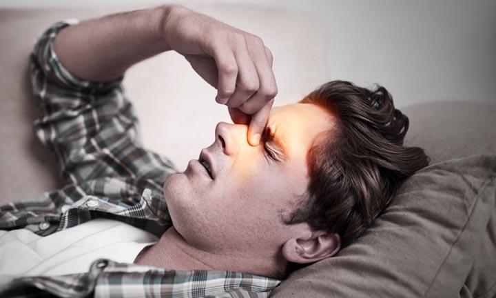 Oberkörpfer und Kopf eines jungen Mannes, der auf dem Sofa liegt und sich aufgrund Sinusitis schmerzverzerrt die Nase masiert
