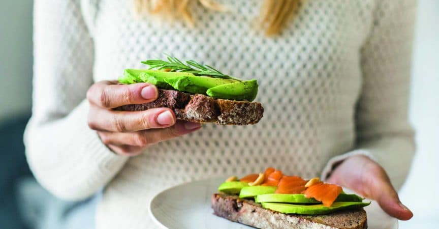Frau hält zwei mit Avocado belegte Brote, eines in der Hand, ein zweites auf einem Teller