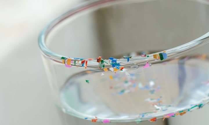 Nahaufnahme des Randes eines gefüllten Wasserglas mit kleinen Plastikteilen am Rand und im Wasser