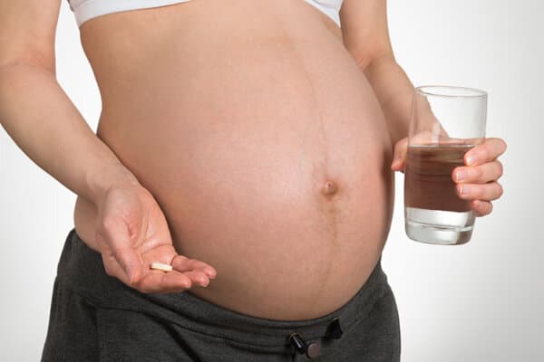 Gros plan d'une femme enceinte tenant un verre d'eau et une pilule de vitamines sur fond gris