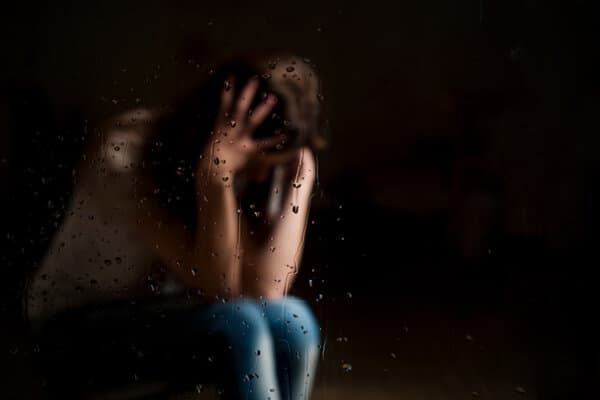 Femme dépressive floue derrière une fenêtre maculée de gouttes de pluie tient sa tête entre ses mains.