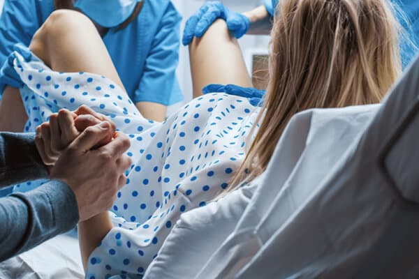 Eine Frau liegt im Geburtssaal auf dem Rücken, vor ihr die Ärzte; ein Mann hält ihre Hand.