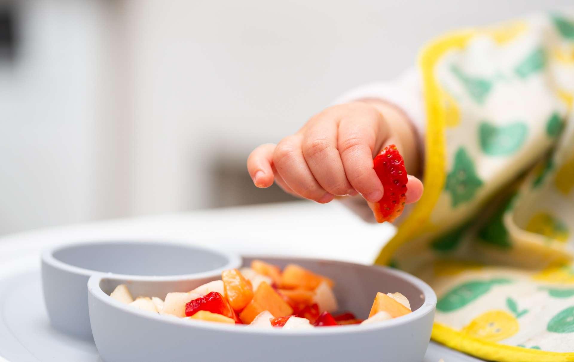 Makro Nahaufnahme der Babyhand mit einem Stück Obst, das im Kinderstuhl sitzt Kind isst gesundes Essen