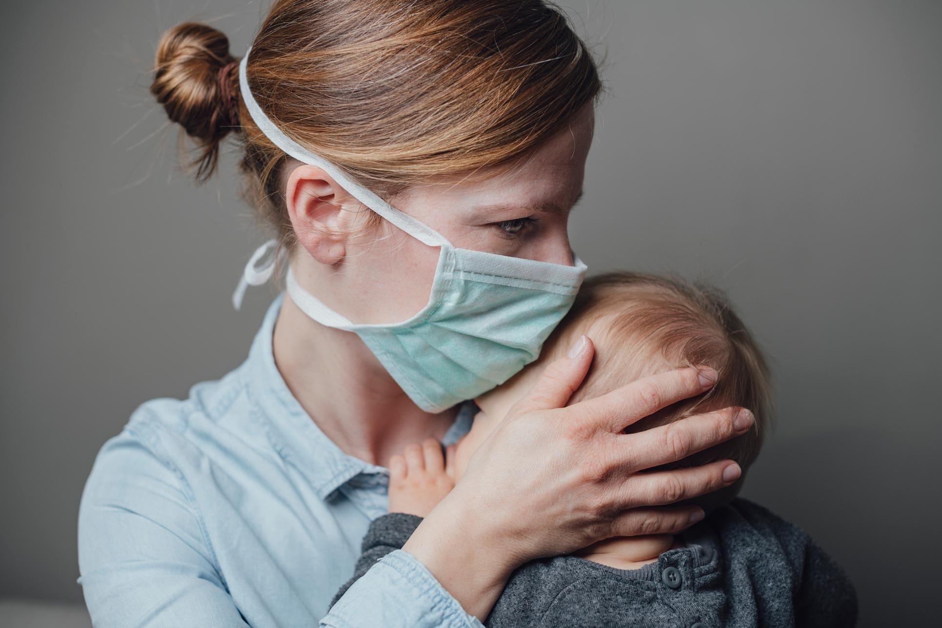 Mutter mit Maske im Gesicht hält ihr Kleinkind in den Armen und mit der Hand stützt sie den Kopf des Kleinkinds