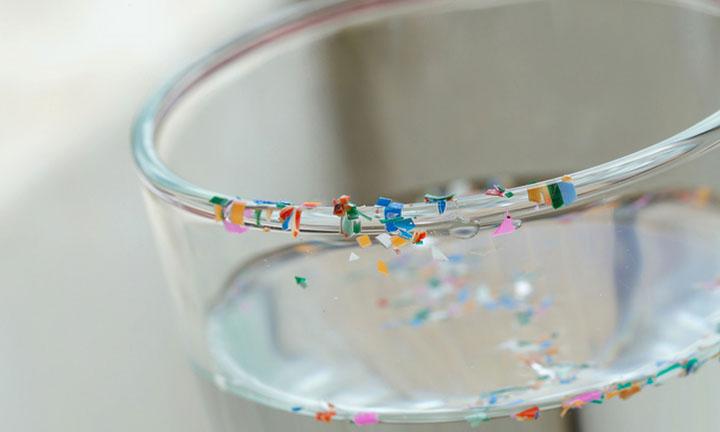Nahaufnahme des Randes eines gefüllten Wasserglas mit kleinen Plastikteilen am Rand und im Wasser