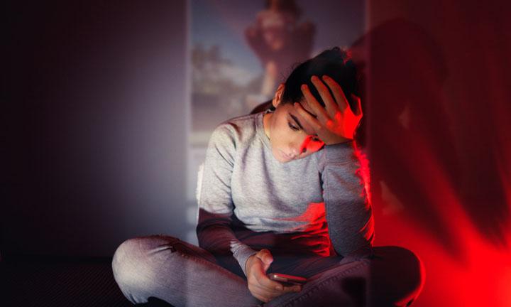 Ein Teenager Mädchen sitzt im Schneidersitz am Boden im Dunkeln vor einer, den Kopf in die eine Hand gestützt, in der anderen hält sie ihr Handy. Ein Projektor projiziert Bilder eines Social Media Feeds auf sie und die Rückwand.