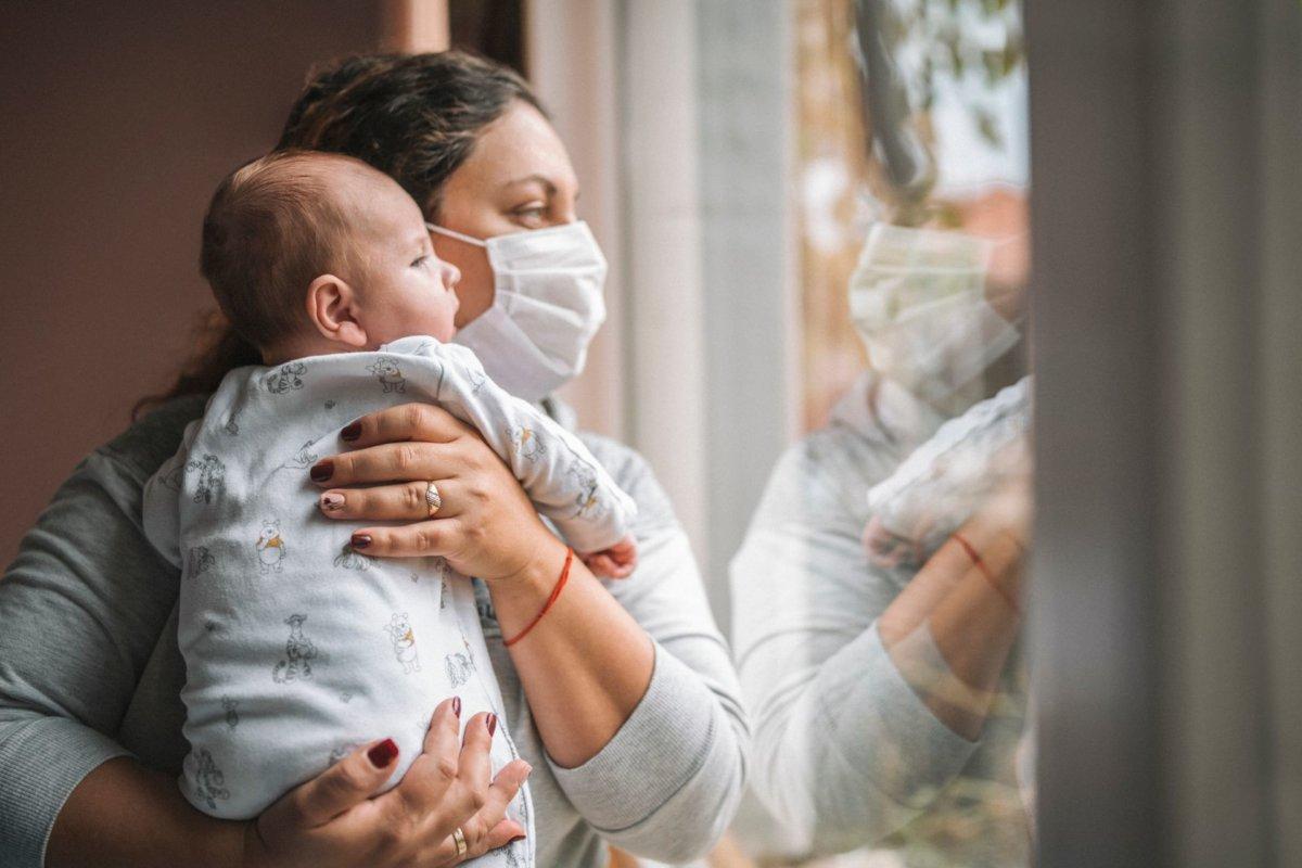 mother with protective face mask holding her newborn baby and they looking trough the window.